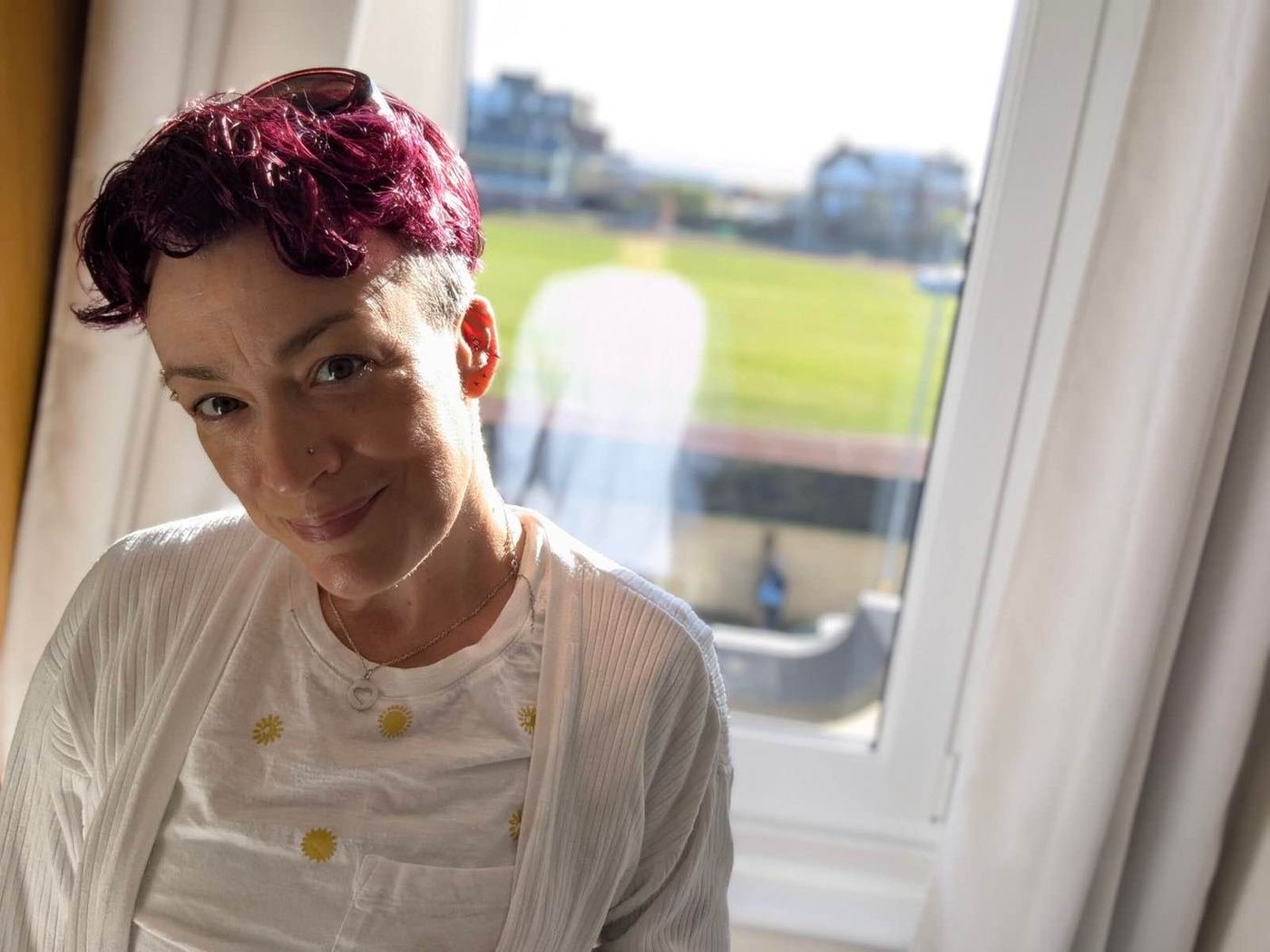 Kate smiling by a sunny window, wearing a white top with yellow buttons