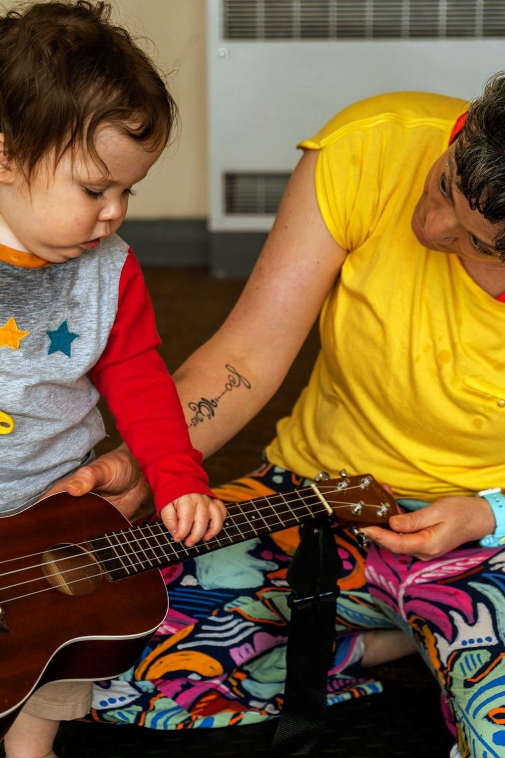Kate, in a yellow top, sitting on the floor beside a toddler who is exploring the strings of her ukulele